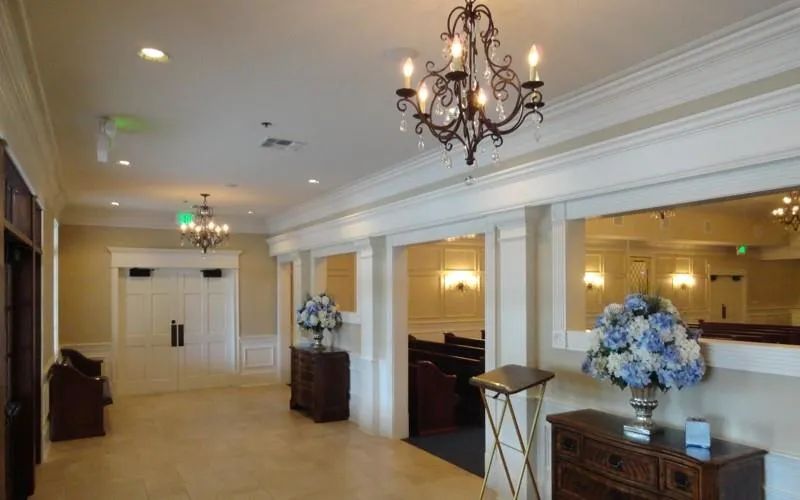 A warm funeral home entryway with a chandelier, floral arrangements, and a guest book stand